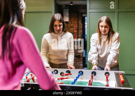 Two young women engaging in a playful match of foosball, with a third friend partially visible, in a bright indoor setting Stock Photo