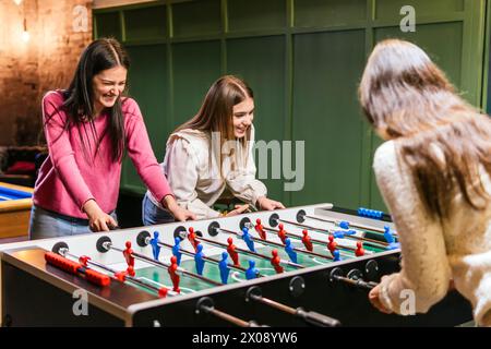 Two young women share a moment of joy and competition, playing foosball in a cozy indoor setting, depicting friendship and leisure time Stock Photo