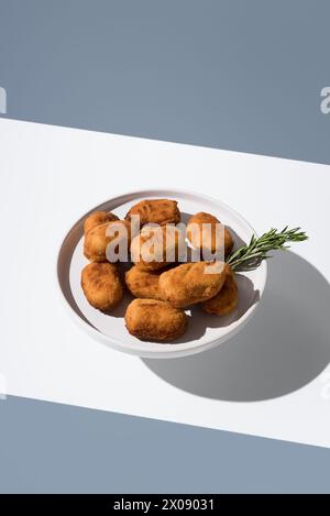 A plate of golden fried croquettes accompanied by a fresh sprig of rosemary, presented on a clean white surface with shadows Stock Photo