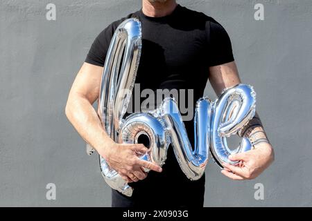 Cropped unrecognizable man in a black t-shirt holds shiny silver balloon letters spelling LOVE, symbolizing a romantic sentiment or celebration. Stock Photo