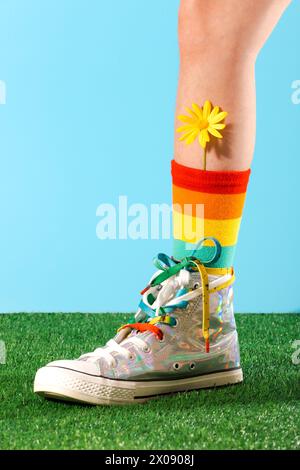 Cropped unrecognizable person wearing vibrant spring-colored socks and sneakers with a yellow flower tucked in, standing on green grass against a blue Stock Photo