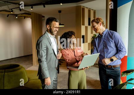 Three colleagues laugh and discuss ideas around a laptop in a vibrant, modern workspace Stock Photo