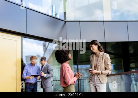 Two female multiethnic colleagues engage in a friendly chat outside an office, with others working in the background Stock Photo