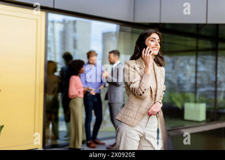 A stylish woman converses on her smartphone, walking past colleagues gathered in casual conversation Stock Photo