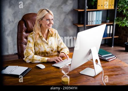 Portrait of successful pretty senior business lady sitting armchair table working looking monitor browsing site workstation workspace Stock Photo