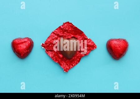 Three red heart-shaped candies on a blue paper background, top view.Two chocolates and one with an unwrapped wrapper. Stock Photo