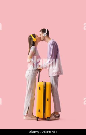 Beautiful young happy couple of tourists in headphones with suitcase kissing on pink background Stock Photo