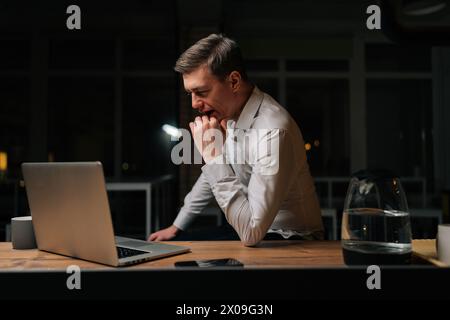 Tired male staff officer yawning during using laptop computer, feeling sleepy after having overwork project overnight. Exhausted businessman working Stock Photo