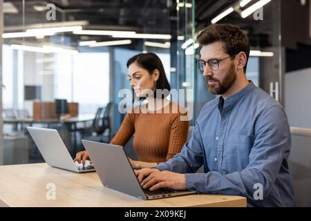 Focused man and woman using laptops at a wooden table in a bright, contemporary workspace, embodying professionalism and teamwork. Stock Photo