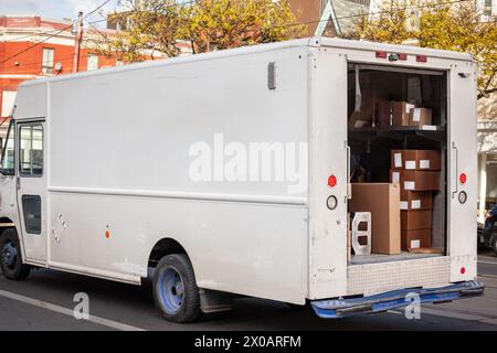 Captured on a city street, this image features a white North American delivery van with its rear doors open, revealing an array of parcels ready for d Stock Photo