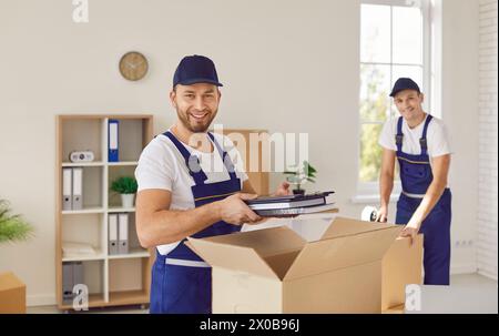Two professional friendly movers from cardboard boxes unpacking office items in new location. Stock Photo