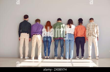 Diverse, silent group of seven people stand in row side by side facing white wall. Stock Photo
