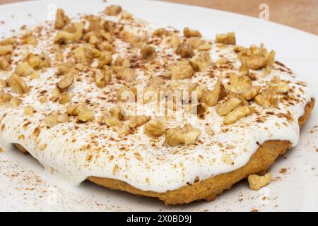 A close-up view of a carrot cake piece, showcasing creamy white chipped cream cheese, crushed nuts, and a hint of cinnamon on a white plate, capturing Stock Photo