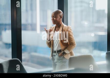 Mature businesswoman in a corporate office standing by a window, holding a smartphone and tablet. She is speaking on the phone while discussing busine Stock Photo
