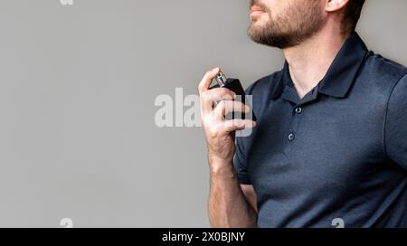 Man in black polo shirt putting on cologne next to light grey wall. Stock Photo