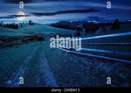 carpathian countryside scenery in spring at night. mountainous rural landscape with path through the meadow and haystack behind the wooden fence in fu Stock Photo