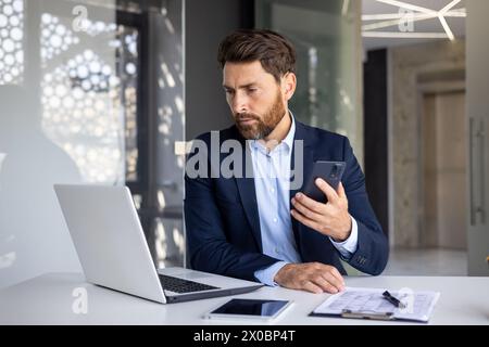 A serious young man businessman sits in the office at the table in a suit, works on a laptop and holds a mobile phone in his hands. Stock Photo