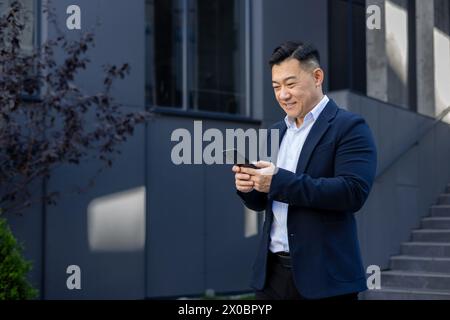 A smiling young Asian male businessman is standing on a city street near a business building, holding and using a mobile phone. Stock Photo