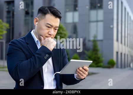 Pensive young Asian male businessman sitting near office building outdoors wearing headphones and looking thoughtfully at tablet screen. Stock Photo
