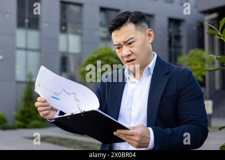 Worried and serious young Asian businessman standing outside office center in suit, holding documents and looking at graphs in frustration. Stock Photo