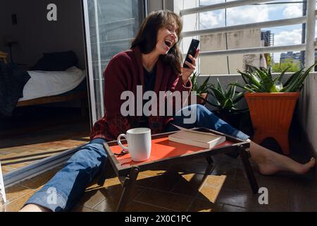 Latin adult woman sitting on balcony of her apartment very happy laughing out loud checking social networks and watching funny videos on phone while r Stock Photo