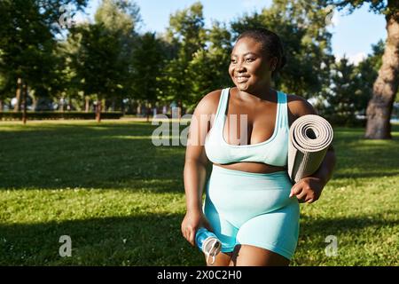 Curvy African American woman in blue sports bra and shorts holding a yoga mat outdoors. Stock Photo