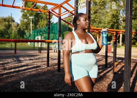 An African American woman in sportswear stands in a park, holding a water bottle, taking a refreshing break from her outdoor workout. Stock Photo