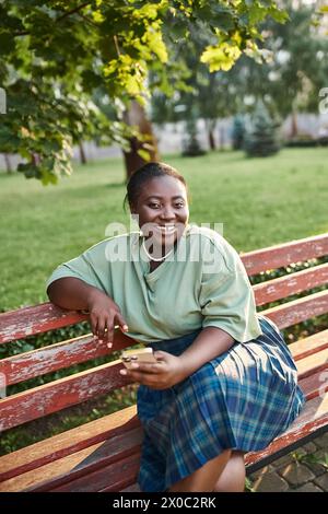Relaxing plus size African American woman sitting on a wooden bench in a peaceful park setting on a sunny day. Stock Photo