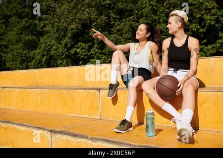 Two young women, athletic friends, sit closely together after playing basketball outdoors on a summer day. Stock Photo