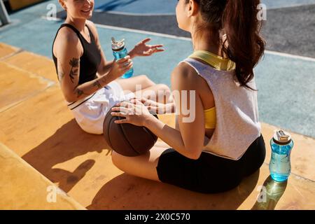 Two young women sit together on the ground, engaged in a deep conversation after playing basketball outdoors in the summer. Stock Photo
