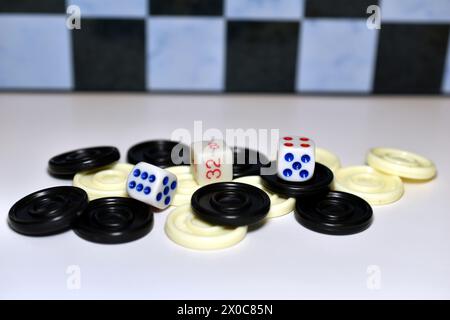 In the picture, on a white table, against the backdrop of a chessboard, dice and chips of two colors stand and lie mixed. Stock Photo