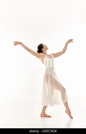 A graceful young woman dances in a flowing white dress against a white studio backdrop. Stock Photo