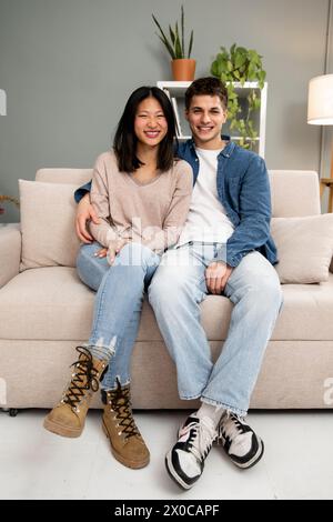 Lovely affectionate smiling young adult couple sitting on sofa at home looking at camera. Diverse carefree man and woman hugging and relaxing together Stock Photo