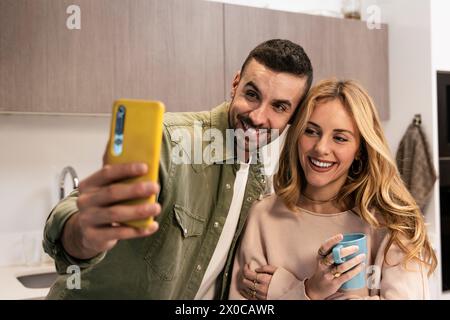 Young joyful couple taking selfie in the kitchen in modern apartment. Young adult man and woman doing a video call while having a drink at home. Stock Photo