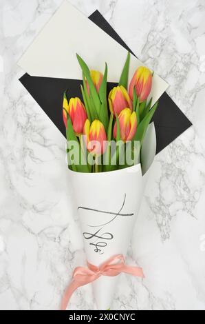 Bouquet of red and yellow flowers with blank sheet of paper and envelope on marble desk. Top view. Stock Photo