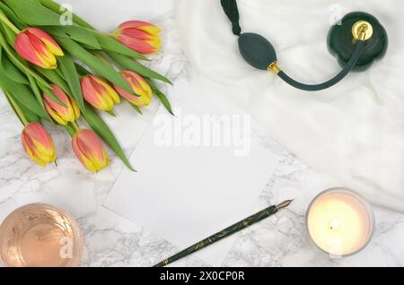 Blank sheet of paper to fill in with content on a table, pen, bunch of tulips, green perfume bottle, candle and rose wine. Stock Photo