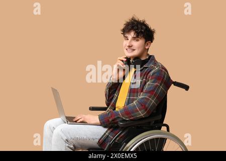 Young male programmer in headphones on wheelchair holding laptop on beige background Stock Photo