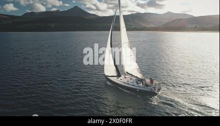 Slow motion yacht sails at mountain island shore aerial. White sailboat at Arran isle, Scotland, Europe. Sun shine above mounts with dark forest at cloudy summer day. Cinematic cruise on boat Stock Photo