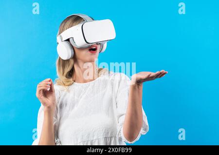 Caucasian girl with VR goggles and standing while holding something and standing at blue background. Woman blowing and looking at hologram in hands Stock Photo