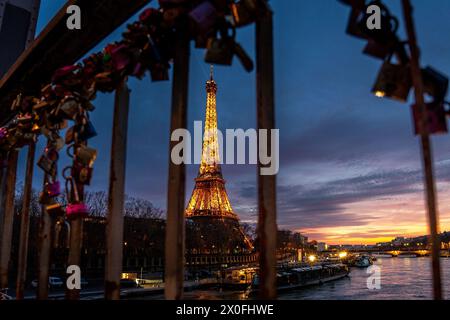The Eiffel Tower seen between the metal bars of a fence filled with love locks shines against the night sky, casting a warm glow on the Parisian city. Stock Photo