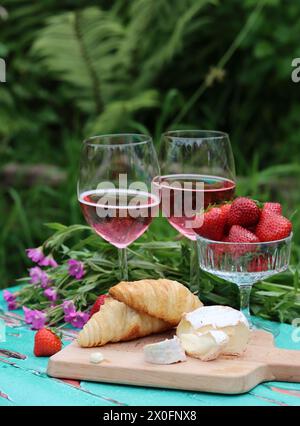 Romantic breakfast in a garden. Summer day weekend. Two glasses of sparkling wine, croissants, strawberry, cheese and pink flowers on a table. Stock Photo
