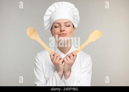 Happy woman chef in uniform holding wooden spoon and spatula on grey background Stock Photo