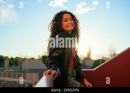 Curly-haired woman smiling on a city rooftop at sunset Stock Photo