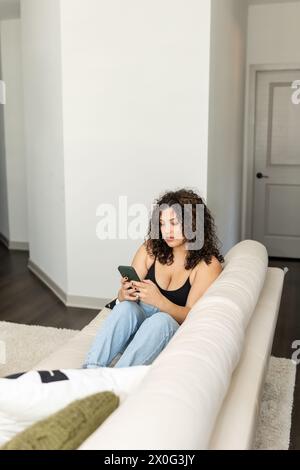 Curly-haired woman immersed in her phone while relaxing on a couch Stock Photo