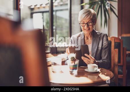 Mature woman is sitting in cafe and relaxing. She is drinking coffee and using digital tablet. Stock Photo