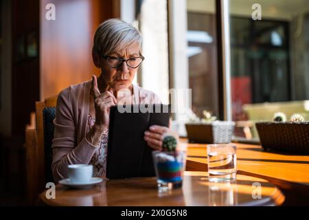 Mature woman is sitting in cafe and using digital tablet. Stock Photo