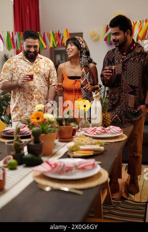 Three cheerful friends in national apparel toasting with glasses of tequila while standing by table with domestic flowers and homemade food Stock Photo