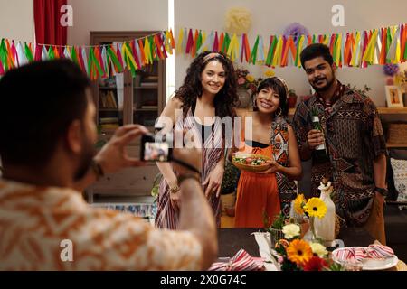 Group of three young cheerful friendly people looking at camera held by their friend taking picture of them during celebration of holiday Stock Photo