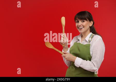 Happy professional confectioner in apron holding wooden spoon and spatula on red background. Space for text Stock Photo