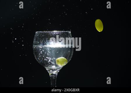 A glass cup with clear water and a grape inside causing splashes. another grape outside the glass, in the air. Stock Photo